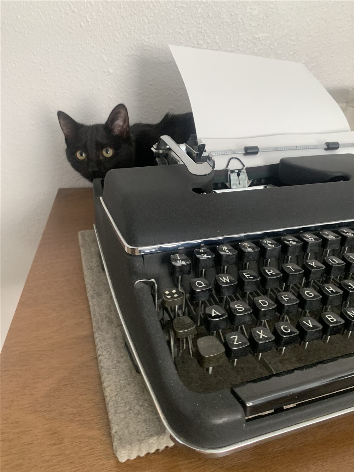 A black cat peeking out from beside a typewriter with a sheet of paper loaded.