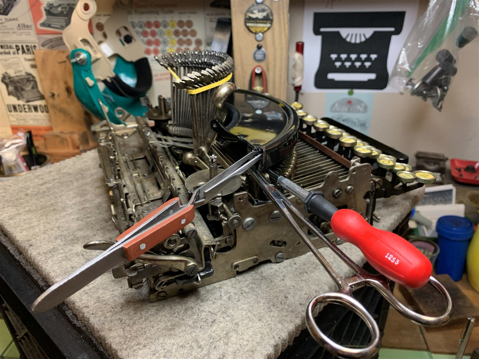 A typewriter carriage and segment mid-teardown on a felt pad, forceps and a screwdriver laid across it, vintage typewriter ads and shop décor on the wall behind.