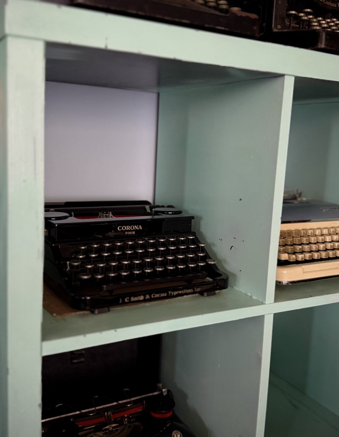A glossy black Corona Four typewriter sitting in a seafoam-green cubby shelf.