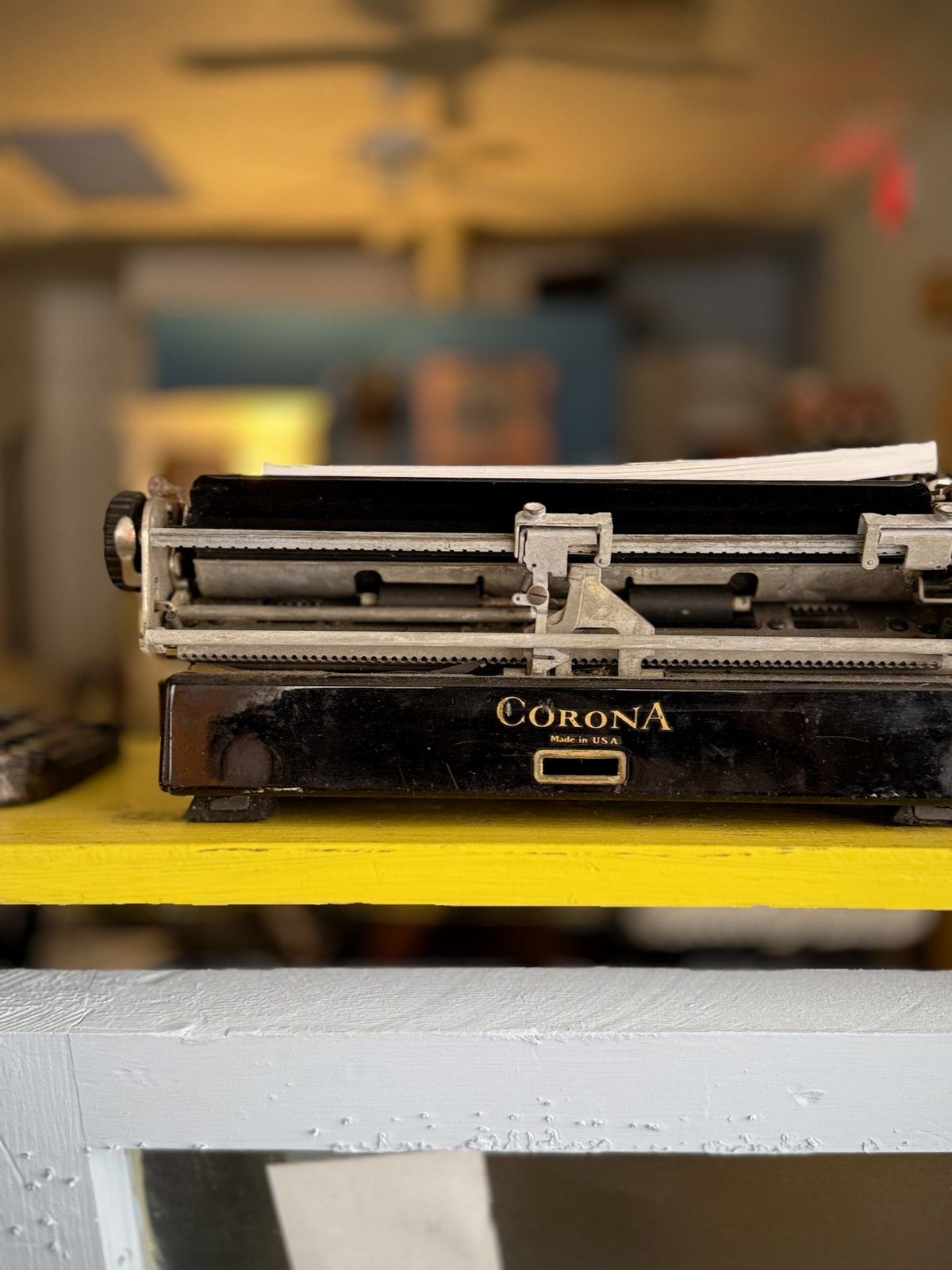 Close-up of a black Corona typewriter's carriage and nameplate, the machine resting on a bright yellow shelf.