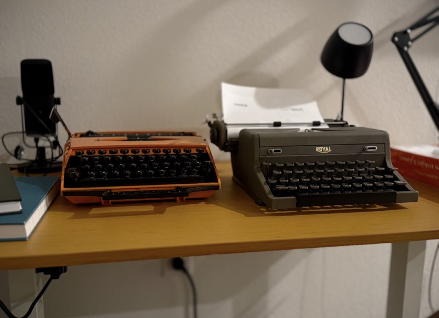 An orange typewriter and a dark gray Royal sit side by side on a desk with a podcast microphone and a stack of books.