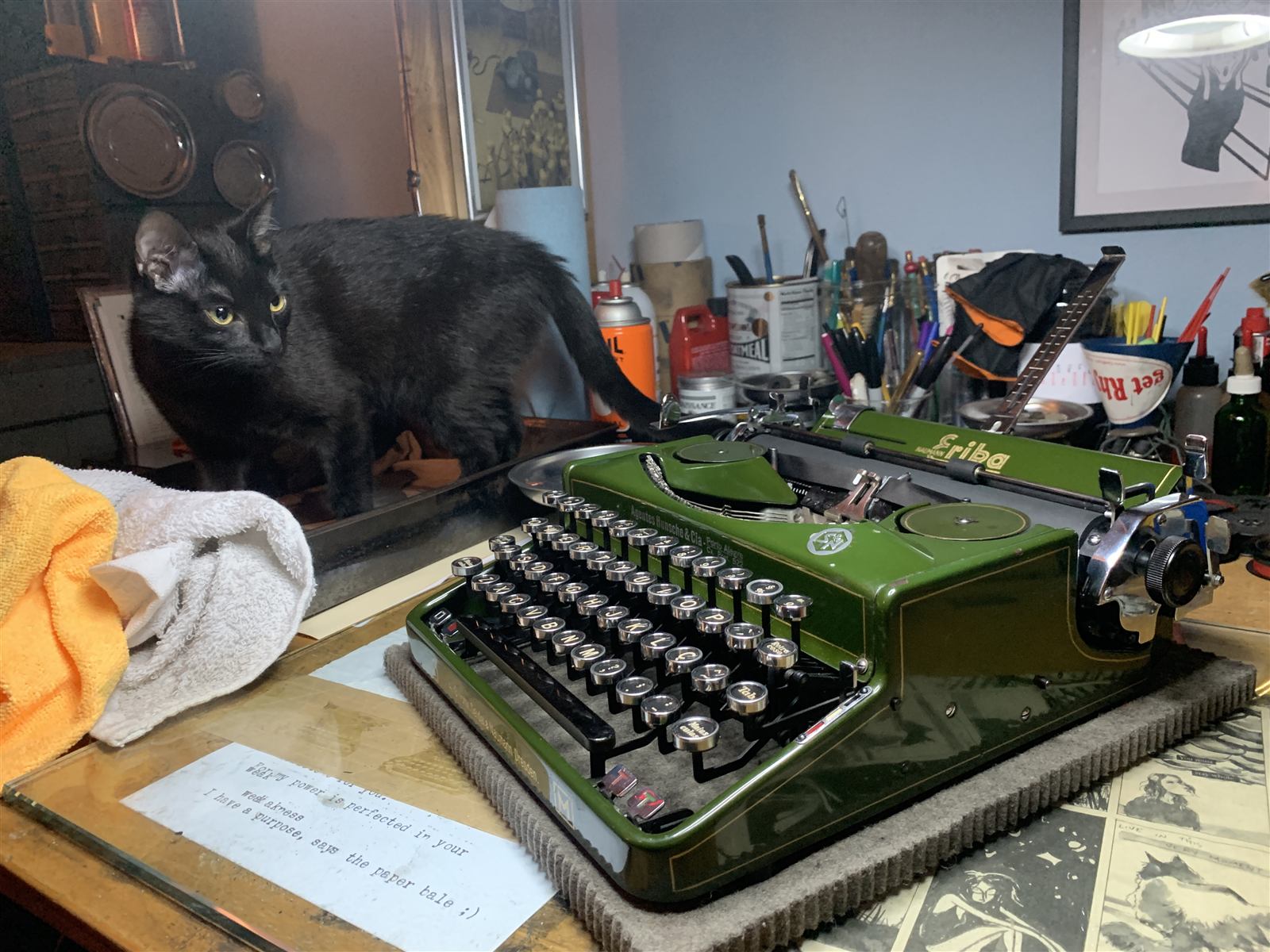 A green Erika typewriter sitting on a desk, with a black cat peering over a shelf just behind it.