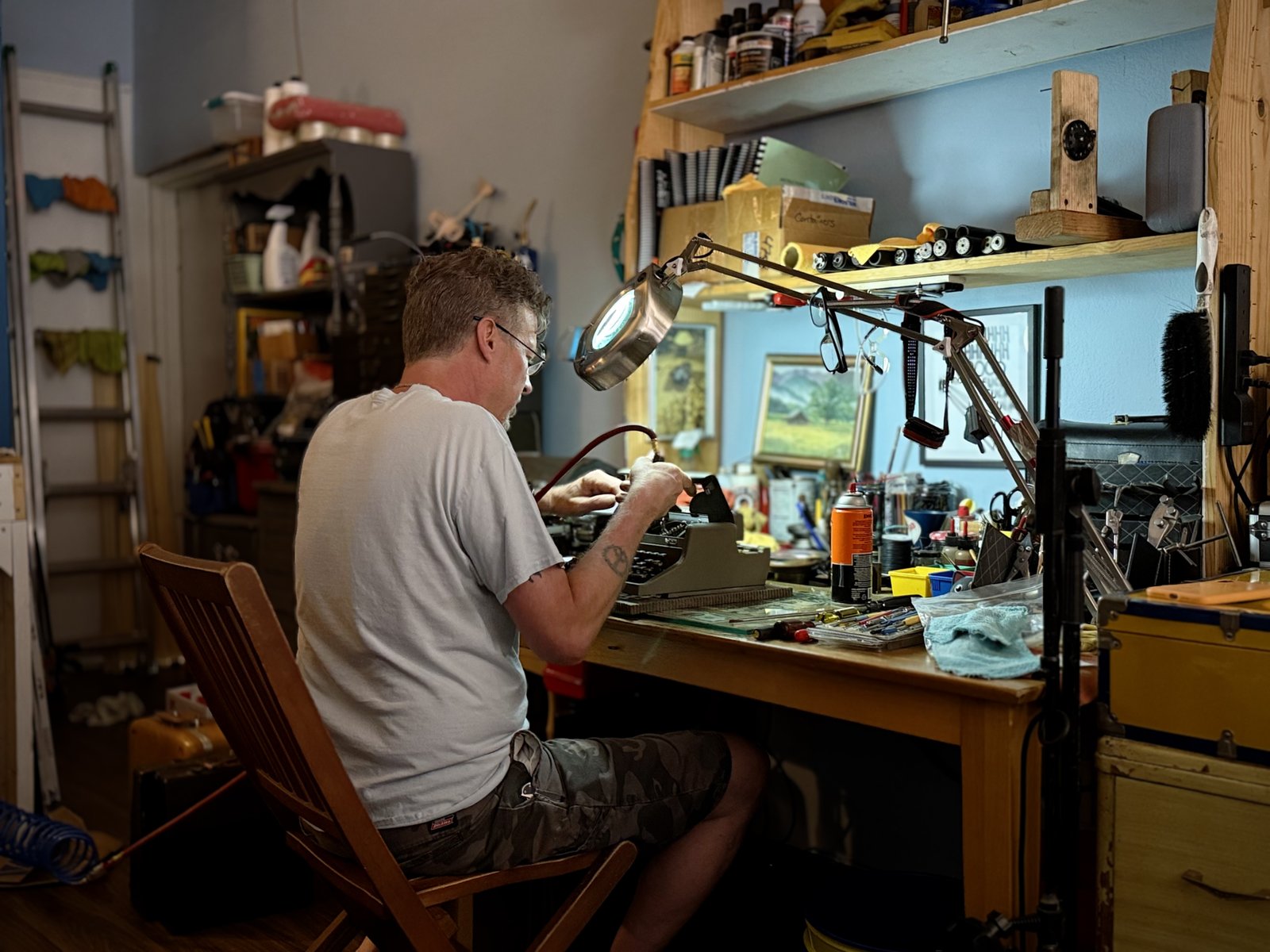 Close-up of hands working on a typewriter's internals at the bench, lamp light catching the tools.