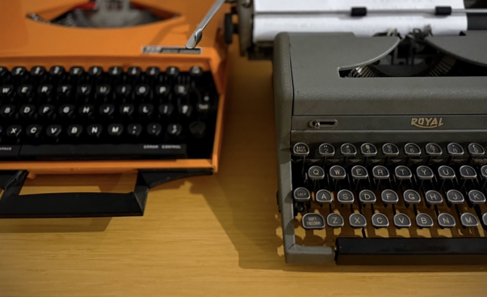 Two portable typewriters side by side on a wooden desk: a bright orange model and a gray Royal with a fresh page rolled into the platen.