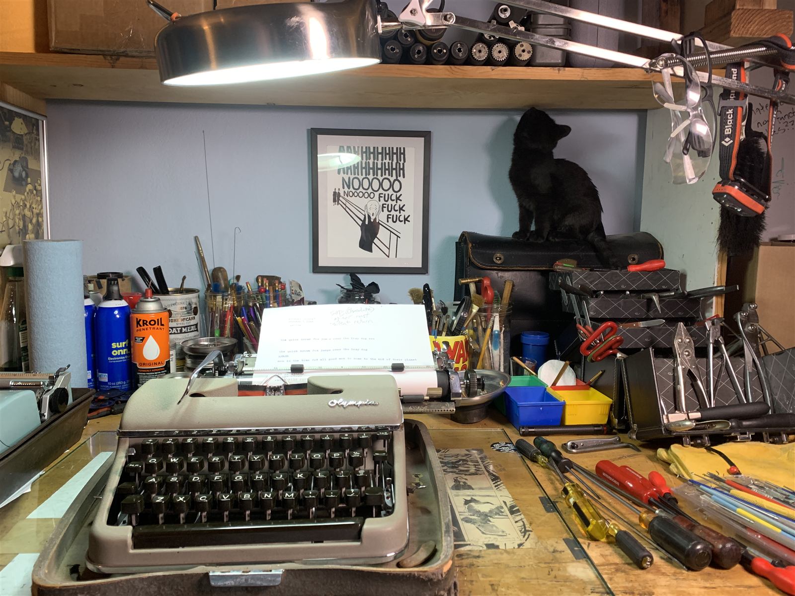 An Olympia typewriter on the workbench, a black cat perched on a toolbox behind it, hand tools laid out alongside.