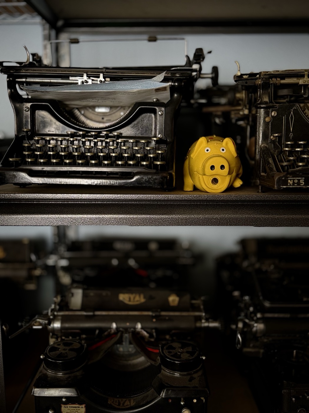 A black vintage typewriter beside a yellow ceramic piggy bank on a shop shelf, another typewriter case marked No. 5 alongside.