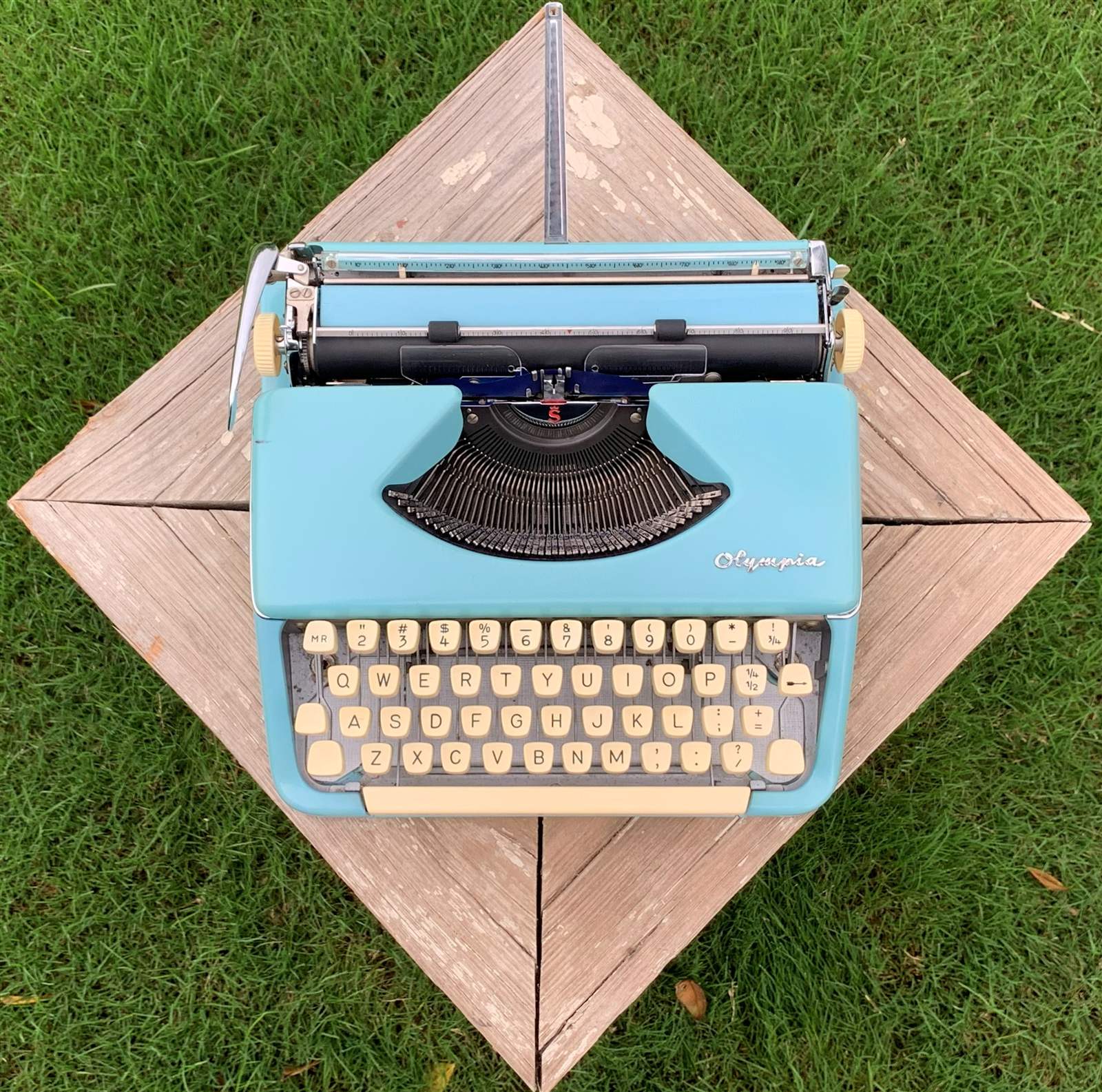 A seafoam-blue Olympia typewriter photographed from directly above, centered on a wood-plank picnic table that reads like a diamond against the grass.