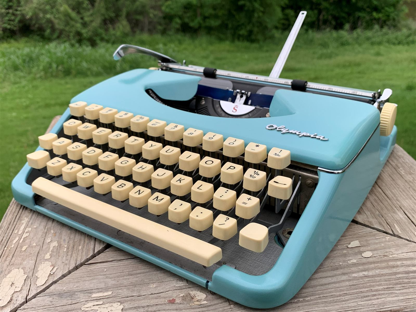 The same seafoam-blue Olympia typewriter from a three-quarter angle on the picnic table, green trees behind.