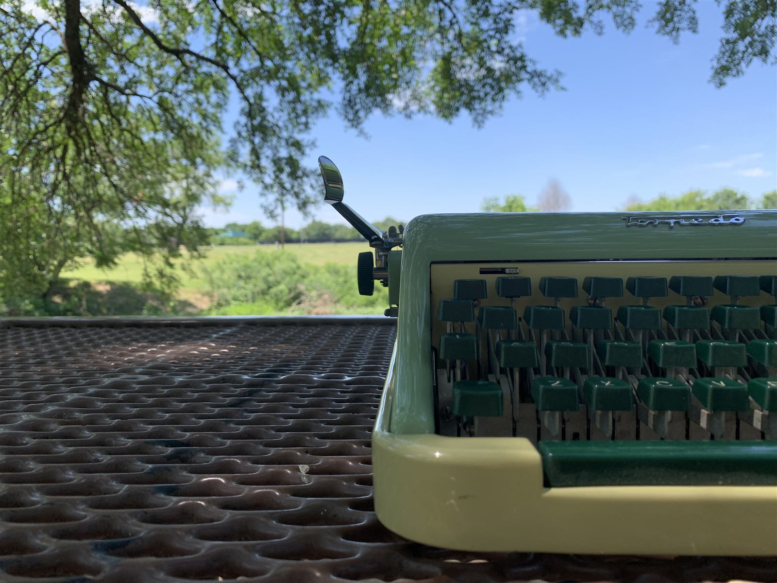 A seafoam-green typewriter on an outdoor table under a tree, blue sky and open field beyond.