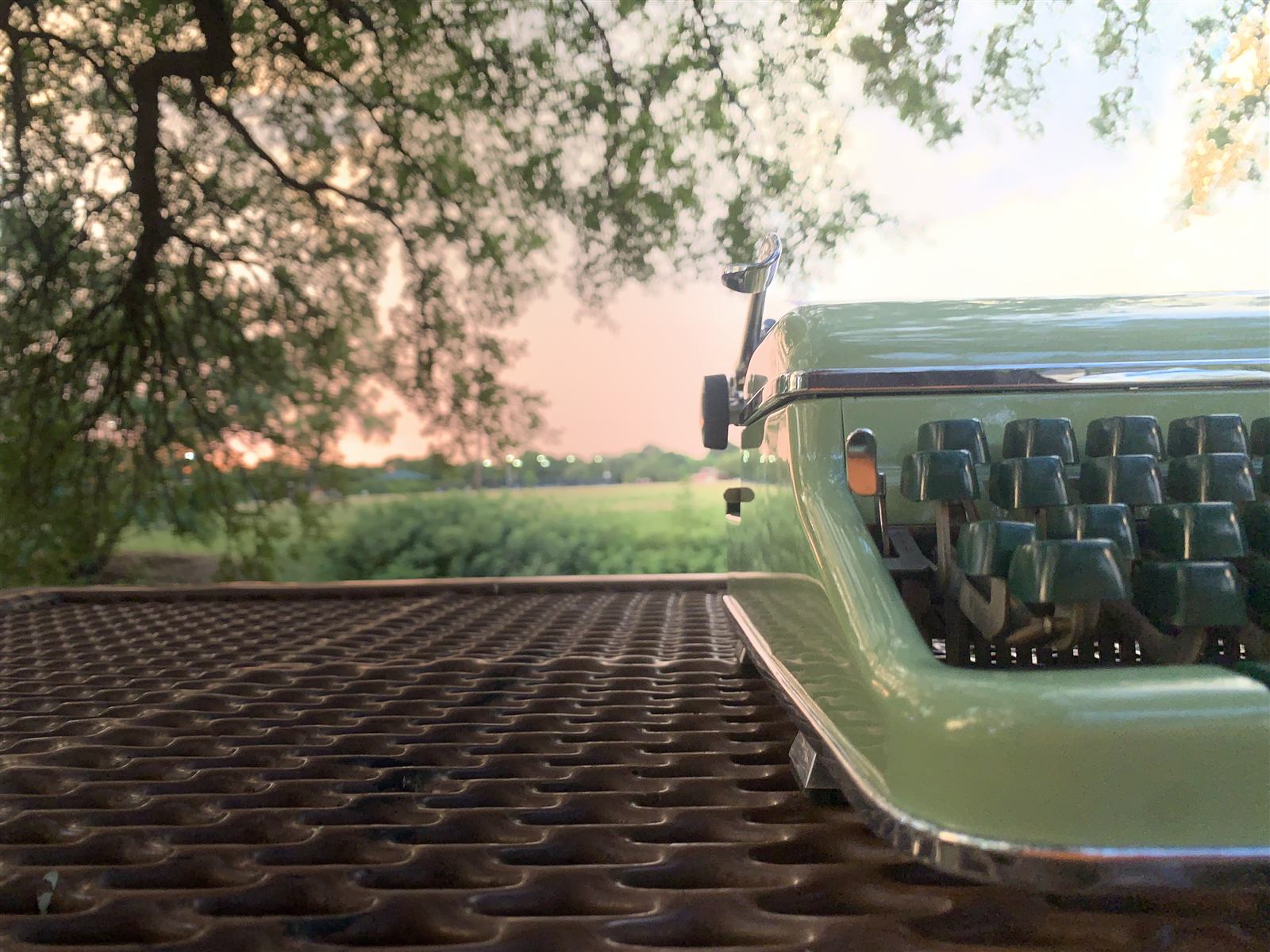 A seafoam-green typewriter on an outdoor table at sunset, pink sky showing through the tree branches.