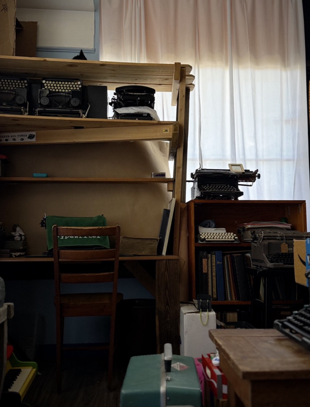 A dim corner of the shop: an empty chair at a wooden shelf-desk, sheer curtains glowing with window light, small typewriters lined up on the shelf above.