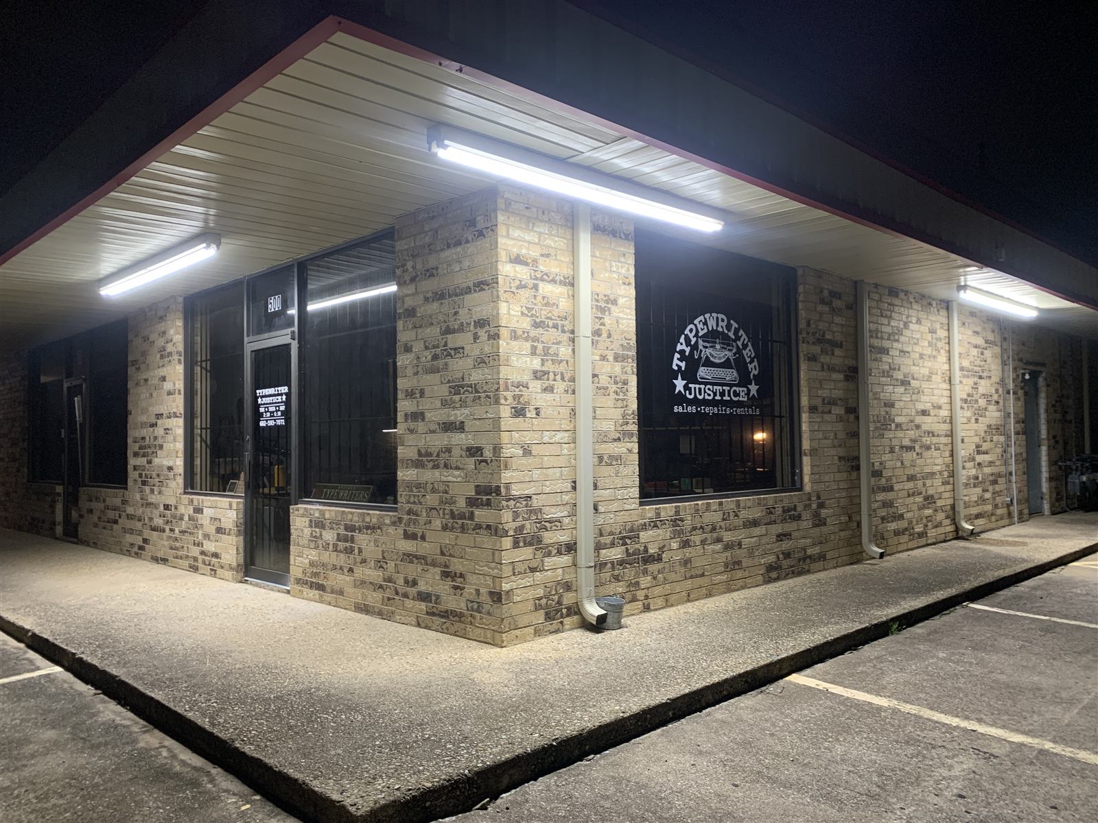 A tight night view of the shop's brick corner, the illuminated Typewriter Justice window sign glowing under the eave lights.