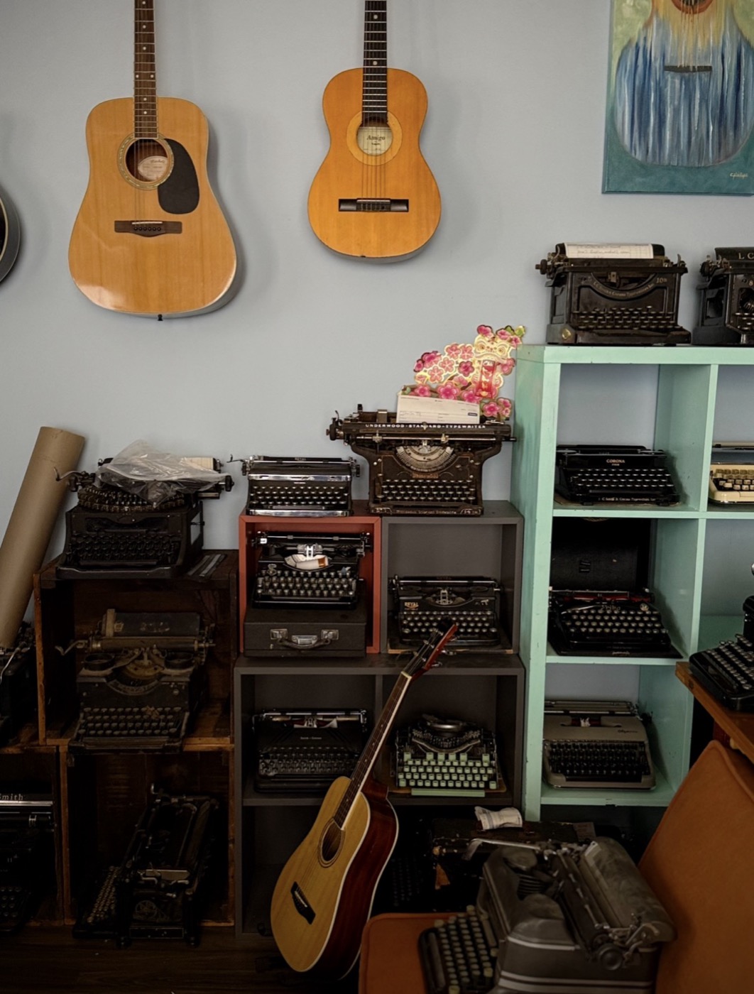 A pale blue shop wall hung with acoustic guitars above shelves stacked with dozens of typewriters of every era and color.