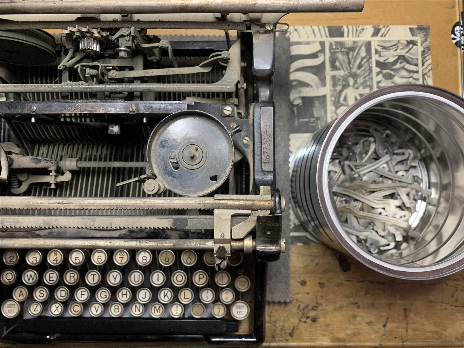 An overhead view of a typewriter opened up mid-teardown on the bench, keyboard exposed, with a tin of typebars alongside.