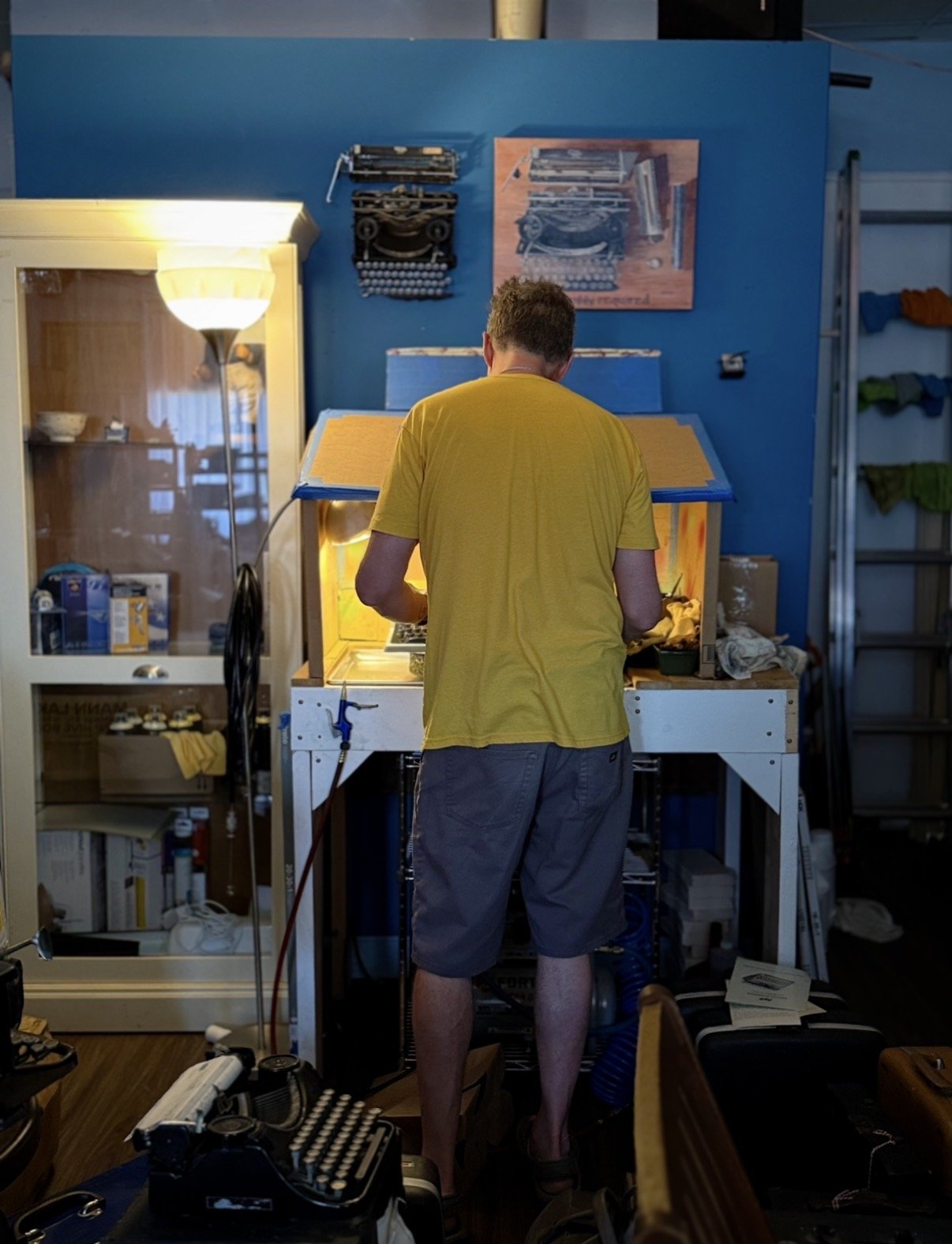 The repairman stands at a blue-walled workbench in the shop, mid-repair on a machine, tools and parts within reach.