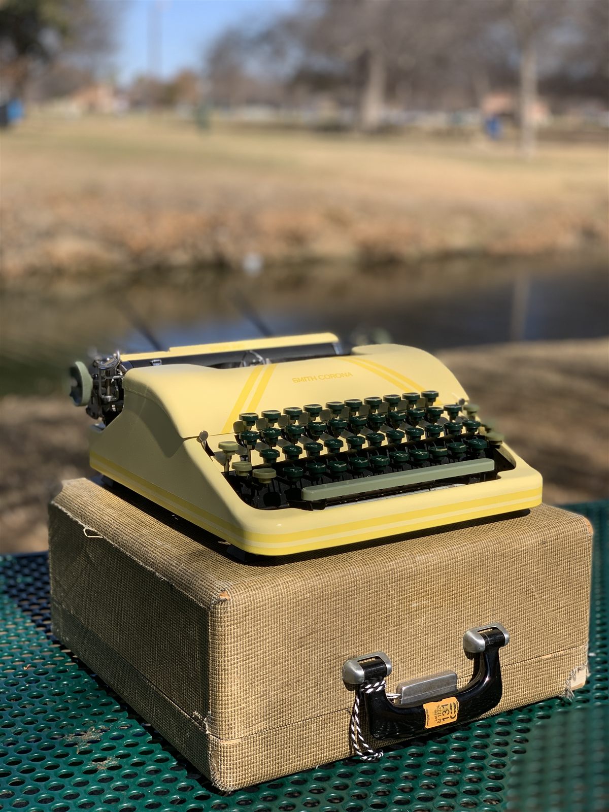 A cream-yellow Smith-Corona typewriter resting on its case outdoors beside a pond.